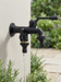 Skade – dual-outlet black outdoor garden faucet on cream stucco wall with water flowing over garden shrubs